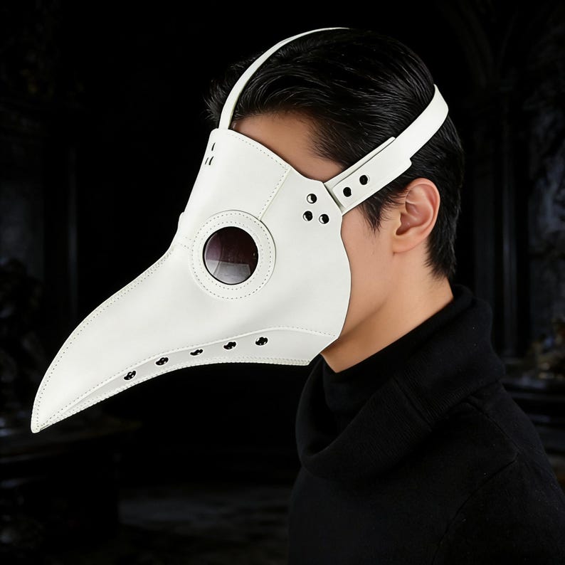 Op de afbeelding: Een witte pestdoktermasker met een lange, gebogen snavel en donkere ooglenzen. Het masker heeft een band die over het hoofd gaat en met klinknagels is bevestigd. Het masker is gemaakt van een glad materiaal.