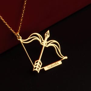 May include: A gold-colored necklace featuring a Sagittarius bow and arrow pendant. The pendant has the name "Isabella" engraved on it. The necklace is displayed against a red and black background.