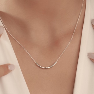 May include: A delicate silver necklace with a series of small, cylindrical beads forming a design. The necklace is worn against a white blouse, with hands gently framing the neckline. The jewelry is simple and elegant.