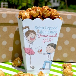 May include: A white popcorn box with a cartoon couple on it. The box says "Brian Popped the Question Janie said YES!" and is filled with caramel popcorn. The box is sitting on a green and white striped surface.