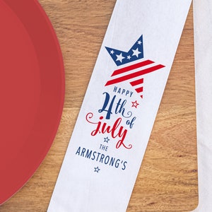 May include: White paper napkin with a red, white, and blue star design. The napkin has the text "Happy 4th of July The Armstrong's" printed on it. A fork, knife, and spoon are tucked into the napkin.