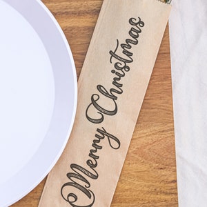 May include: A brown paper napkin with the text "Merry Christmas" printed on it. A set of silverware is wrapped inside the napkin.