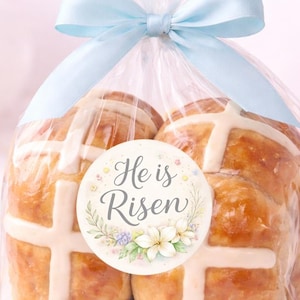 May include: A clear cellophane bag tied with a light blue ribbon, containing several golden-brown hot cross buns. Each bun has a white cross on top. A round sticker with the words "He is Risen" and floral accents is on the front.