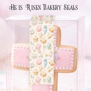 May include: A cross-shaped cookie with pink icing and pearl-like decorations, packaged in a clear box. The cookie is decorated with a patterned sticker that reads "He is Risen." The packaging includes the text "High Gloss Sticker Strips 5-1/2" x 3-1/3" Each."
