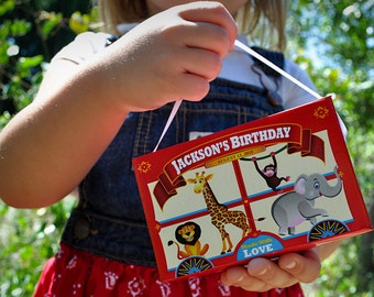 May include: A red, white, and blue circus-themed birthday party favor box with the text "Jackson's Birthday" and "Made with Love". The box features illustrations of a giraffe, elephant, lion, and monkey.