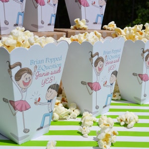 May include: White popcorn boxes with a blue and pink cartoon illustration of a couple getting engaged. The text on the boxes reads "Brian Popped the Question Janie said YES!"