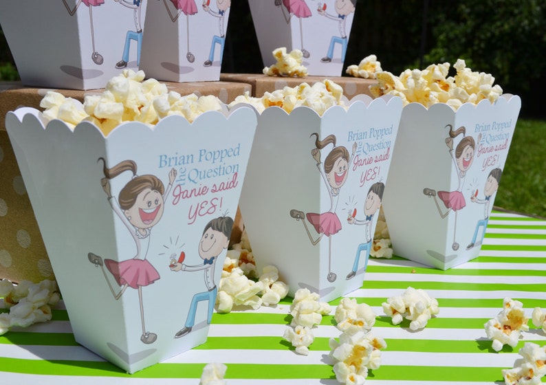 May include: White popcorn boxes with a blue and pink cartoon illustration of a couple getting engaged. The text on the boxes reads "Brian Popped the Question Janie said YES!"