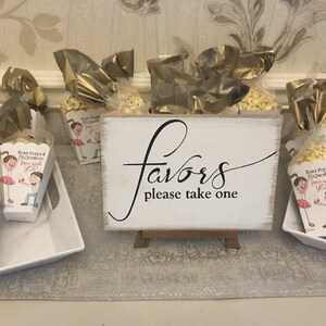 May include: A white sign with black calligraphy that reads "Favors please take one". The sign is on a small wooden easel. In front of the sign are small white boxes filled with popcorn and decorated with a cartoon couple. The boxes are wrapped in gold cellophane.