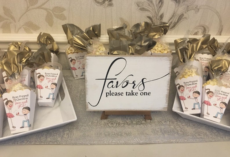 May include: A white sign with black calligraphy that reads "Favors please take one". The sign is on a small wooden easel. In front of the sign are small white boxes filled with popcorn and decorated with a cartoon couple. The boxes are wrapped in gold cellophane.