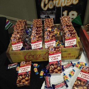 May include: A wooden Coca-Cola crate filled with boxes of treats labeled "Congrats Caleb." Assorted candies and Baby Ruth bars are scattered around the crate. A baseball jersey with the word "Corner" is in the background.
