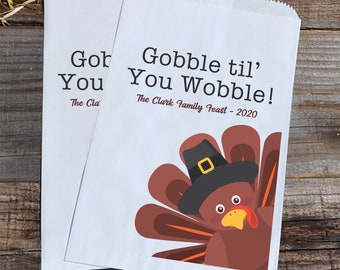 May include: Two white paper bags with a Thanksgiving turkey design. The text on the bag reads "Gobble til' You Wobble! The Clark Family Feast - 2020".
