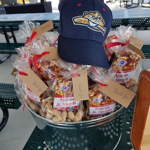 May include: A navy blue baseball cap with a team logo sits atop a metal bucket filled with peanuts and wrapped snacks. The snacks are labeled "Norm & Steph" and tied with red ribbons. "Thank You" tags are attached.