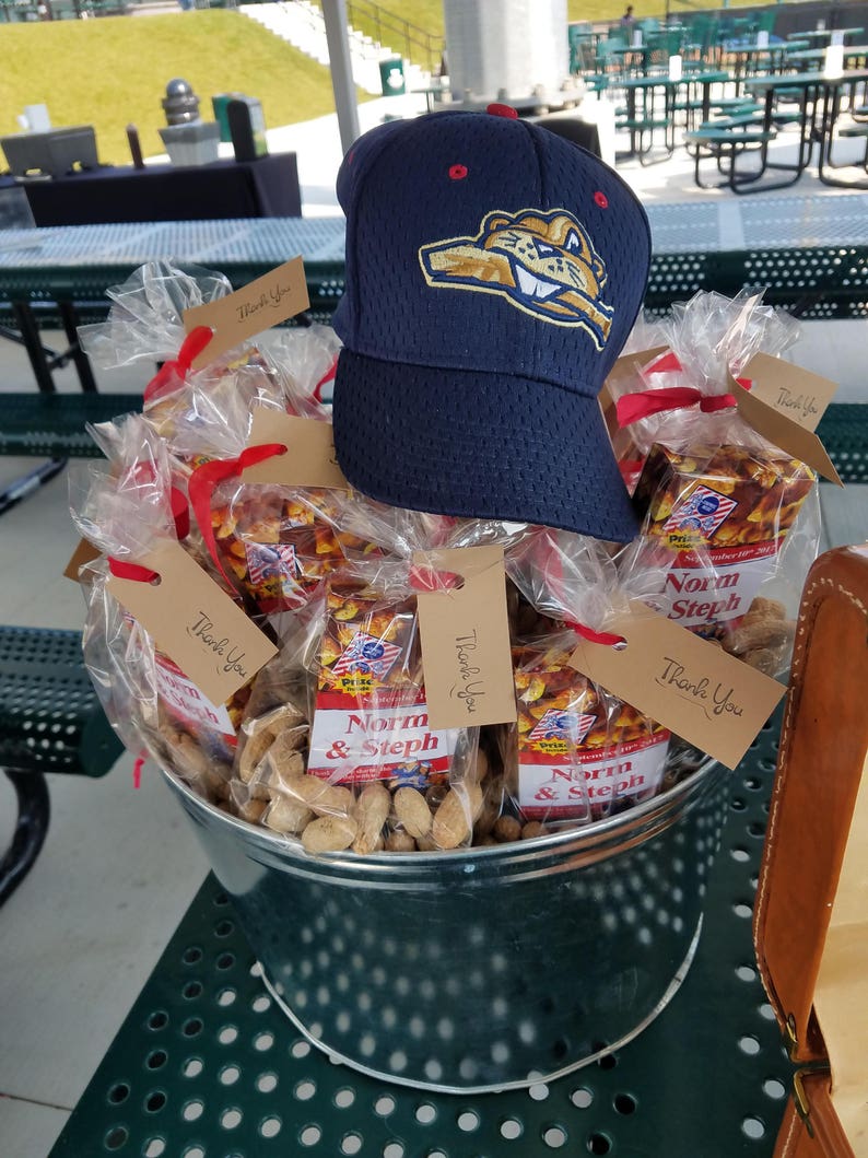 May include: A navy blue baseball cap with a team logo sits atop a metal bucket filled with peanuts and wrapped snacks. The snacks are labeled "Norm & Steph" and tied with red ribbons. "Thank You" tags are attached.