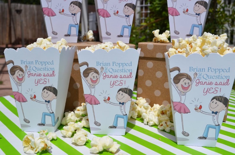 May include: Popcorn boxes with a blue and white design featuring a cartoon couple. The boxes have the text "Brian Popped the Question Janie said YES!" printed on them.