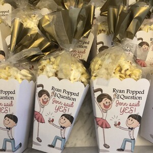 May include: White popcorn boxes with a cartoon illustration of a couple getting engaged. The boxes have the text "Ryan Popped the Question Jenn said YES!" printed on them.