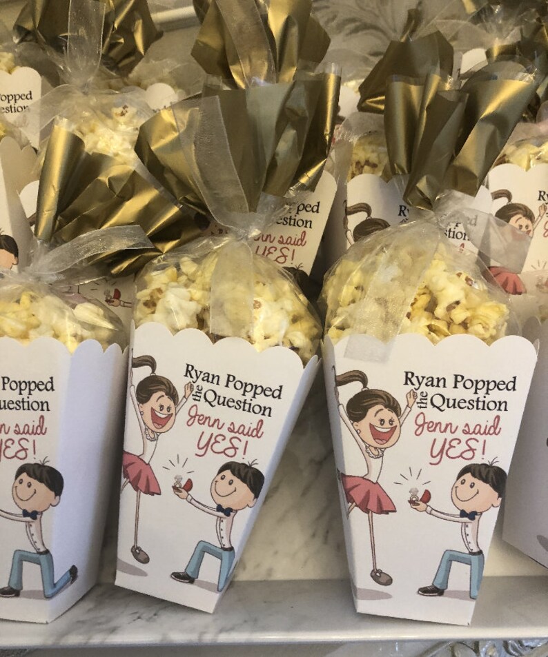 May include: White popcorn boxes with a cartoon illustration of a couple getting engaged. The boxes have the text "Ryan Popped the Question Jenn said YES!" printed on them.
