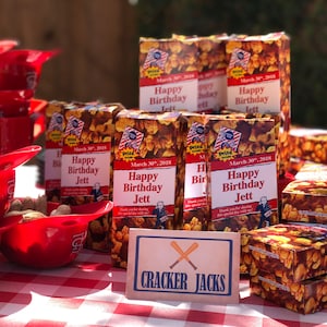 May include: A festive display of Cracker Jacks boxes and red baseball-themed bowls filled with peanuts. The boxes are personalized with "Happy Birthday Jett" and the date March 30, 2018. A sign reads "Cracker Jacks."