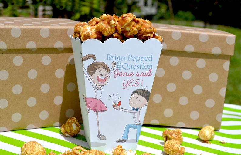 May include: A white popcorn box with a cartoon couple on it. The box says "Brian Popped the Question Janie said YES!" and is filled with caramel popcorn. The box is sitting on a green and white striped surface.