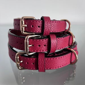May include: A stack of three magenta textured leather collars, each featuring a polished rose gold metal buckle, a matching metal loop, and a fixed leather keeper. The collars have visible stitching along the edges and multiple adjustment holes.