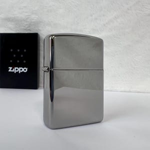 Pode incluir: Um isqueiro Zippo cinza metálico brilhante, de formato retangular. O isqueiro está fechado, com uma dobradiça visível e uma superfície lisa e reflexiva. Uma caixa preta com o logotipo Zippo está ao fundo.