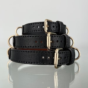 Puede incluir: Tres cinturones negros apilados con un patrón texturizado de piel de lagarto y hebillas de metal en tono dorado. Cada cinturón presenta una serie de agujeros perforados para el ajuste y un anillo de metal circular en el lateral, con diseño uniforme.