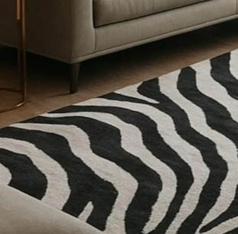 May include: A black and white zebra-striped area rug. The rug is rectangular and features a repeating pattern of black stripes on a light background. The rug is placed on a wooden floor, with a gray sofa visible in the background.