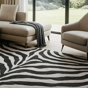 May include: A black and white zebra-striped area rug in a living room setting. The rug is placed in front of a beige sectional sofa and armchair. A dark gray throw blanket is draped over the sofa. Large windows provide a view of the outdoors.