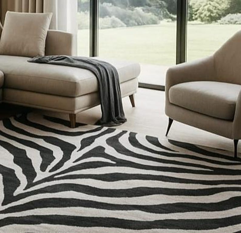 May include: A black and white zebra-striped area rug in a living room setting. The rug is placed in front of a beige sectional sofa and armchair. A dark gray throw blanket is draped over the sofa. Large windows provide a view of the outdoors.