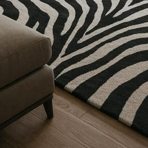 May include: A close-up of a zebra-striped rug in black and beige, with a section of a brown upholstered sofa in the foreground. The rug's pattern is bold and graphic, contrasting with the wooden floor.