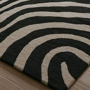 May include: A close-up of a rug with a zebra stripe pattern. The rug features alternating black and beige curved stripes. The rug is made of a soft, textured material and is placed on a wooden floor.