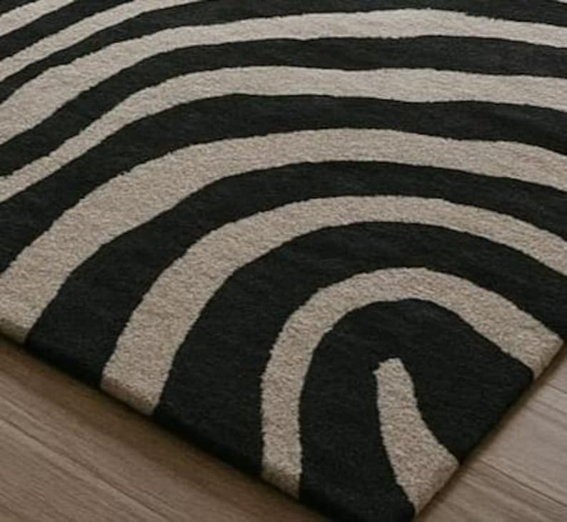 May include: A close-up of a rug with a zebra stripe pattern. The rug features alternating black and beige curved stripes. The rug is made of a soft, textured material and is placed on a wooden floor.