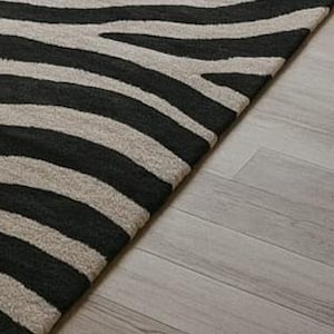 May include: A black and beige zebra-striped area rug. The rug's pattern features bold, dark stripes against a lighter background. The rug is placed on a light-colored wooden floor, with a portion of the floor visible.