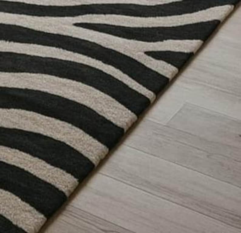 May include: A black and beige zebra-striped area rug. The rug's pattern features bold, dark stripes against a lighter background. The rug is placed on a light-colored wooden floor, with a portion of the floor visible.