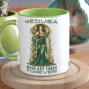 Peut inclure: Mug en céramique blanche avec intérieur et anse verts. L'illustration de Méduse en robe verte est imprimée sur le devant, avec les textes « MEDUSA » et « WASH DAY TAKES FOREVER ! ». Le mug est posé sur un dessous de verre en bois.