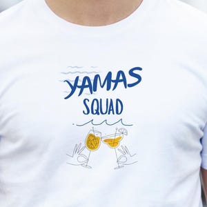Puede incluir: Camiseta blanca con la frase "YAMAS SQUAD" en azul sobre un dibujo lineal de dos cócteles. El diseño incluye una ola. La camiseta es de cuello redondo.