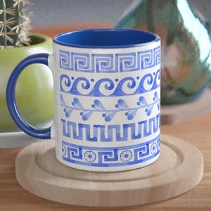 Puede incluir: Una taza de cerámica blanca con interior y asa azules, decorada con patrones azules de grecas y olas. La taza está sobre un posavasos de madera, con un pequeño cactus en una maceta verde y un jarrón turquesa al fondo.