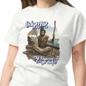 May include: White t-shirt featuring a graphic of Odysseus on a ship, holding a scroll and staff. The text "Odysseus" is above the image, with "Still figuring out Google Maps!" below.