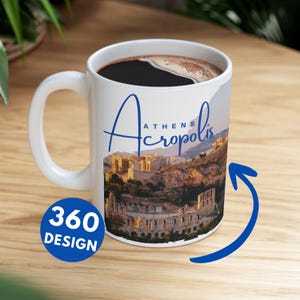 Op de afbeelding: Witte keramische koffiemok met een schilderachtig uitzicht op de Akropolis van Athene. De mok heeft een gebogen handvat en is gevuld met donkere koffie. De woorden "Athens Acropolis" zijn in blauw geschreven. Een blauwe 360 design sticker is op de mok.