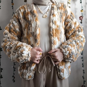 Op de afbeelding: Chunky gebreid vest in crème, bruin en grijs. De open voorkant trui heeft een getextureerd, oversized ontwerp. De mouwen zijn lang en volumineus. Het vest wordt gedragen over een crèmekleurige top en een bruine broek.