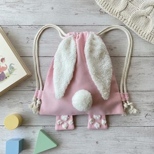 Op de afbeelding: Een roze rugzak met trekkoord, ontworpen als een konijn, met witte, pluizige oren, een pomponstaart en geborduurde pootafdrukken. Het heeft crèmekleurige trekkoorden. Een kinderaccessoire.