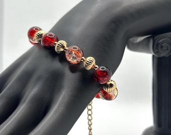 May include: A beaded bracelet featuring alternating red and gold-colored beads. The red beads have a crackled appearance, and the gold beads have a textured, ribbed design. The bracelet is displayed on a black hand.