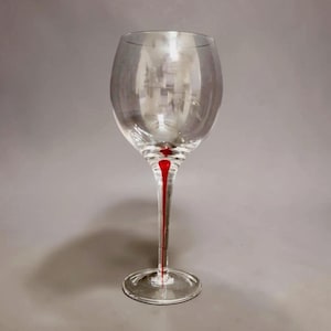 May include: A clear glass wine goblet featuring a rounded bowl and a slender stem. A single, vertical, vibrant red line runs down the center of the stem, extending from the base of the bowl to the circular foot of the glass.