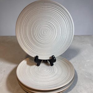 May include: A set of four off-white ceramic plates with a spiral design. The plates are stacked, with one plate displayed on a black stand. The plates have a speckled texture and a slightly raised rim.