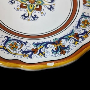May include: Close-up of a decorative ceramic plate with a white center and a brown border. The plate features intricate floral patterns in blue, yellow, and orange. The rim has a scalloped edge with a yellow and orange trim.