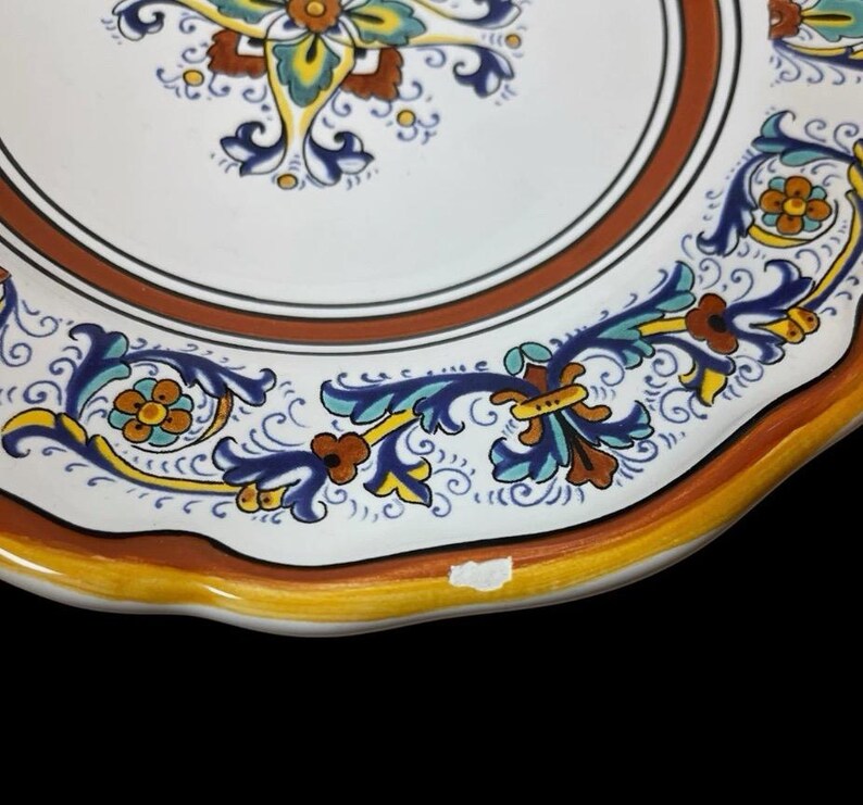 May include: Close-up of a decorative ceramic plate with a white center and a brown border. The plate features intricate floral patterns in blue, yellow, and orange. The rim has a scalloped edge with a yellow and orange trim.