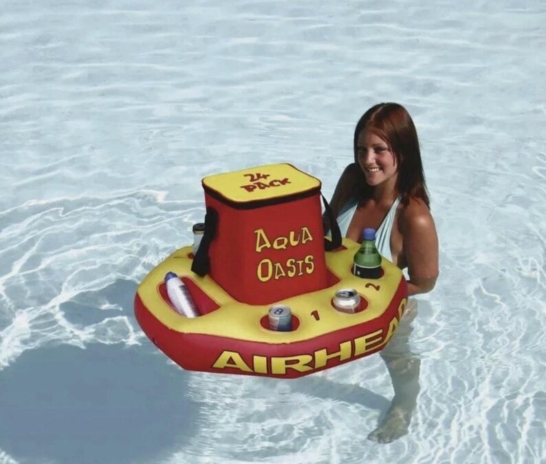 Puede incluir: Un enfriador flotante inflable rojo y amarillo con las palabras "Aqua Oasis" y "Airhead" impresas. El enfriador tiene una parte superior amarilla con las palabras "24 Pack" y contiene latas y una botella. El enfriador est&aacute; en una piscina.