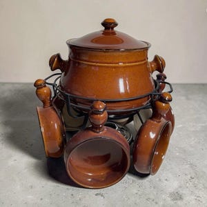 Puede incluir: Un juego de fondue de cerámica marrón con tapa a juego, asas y seis cuencos pequeños. El juego está soportado por un soporte de metal negro. La olla de fondue y los cuencos tienen un acabado brillante.