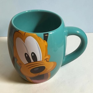 May include: A teal ceramic mug featuring a large, colorful graphic of the cartoon character Pluto on the front. The mug has a rounded, bulbous shape with a sturdy handle on the right side and a smooth, glossy finish throughout the exterior surface.