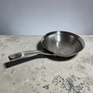 May include: Stainless steel frying pan with a long handle. The pan is round with a slightly sloped edge. The handle has a loop at the end for hanging. The pan is sitting on a concrete surface.
