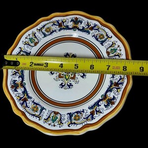 May include: A decorative ceramic plate with a scalloped edge. The plate features a white center with a brown border and intricate floral designs in blue, green, and yellow. The plate measures approximately 8 inches in diameter.